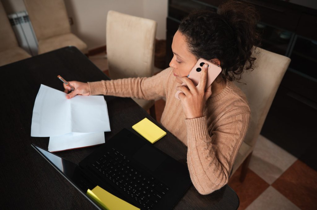 A woman efficiently manages her tasks by handling paperwork, a phone call, and using her laptop.