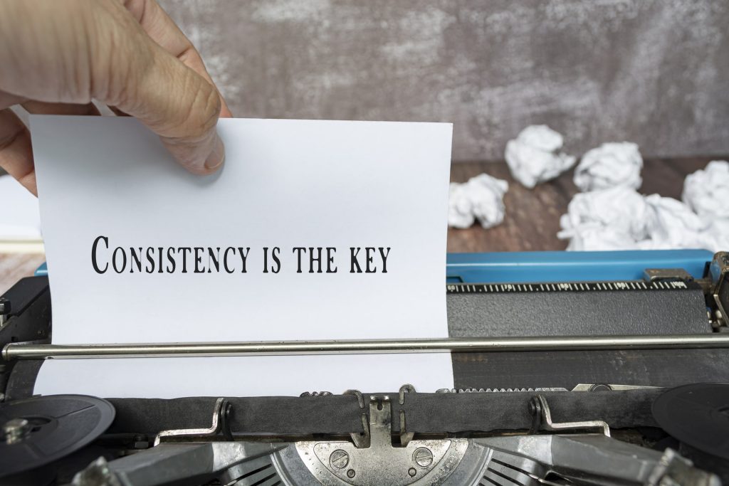 Hand pulling a piece of white paper with text from an old classic typewriter on wooden desk - Consistency is the key.