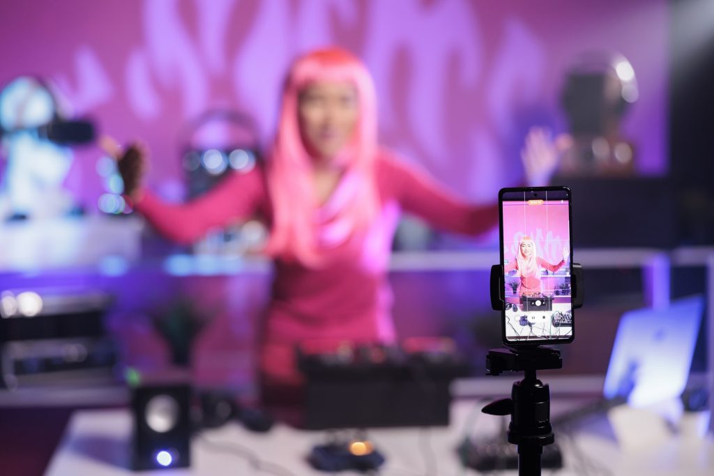 Woman with pink hair standing at dj table mixing song with turntables in front of camera while recording video with phone. Smiling artist doing performance at nightclub with audio equipment.