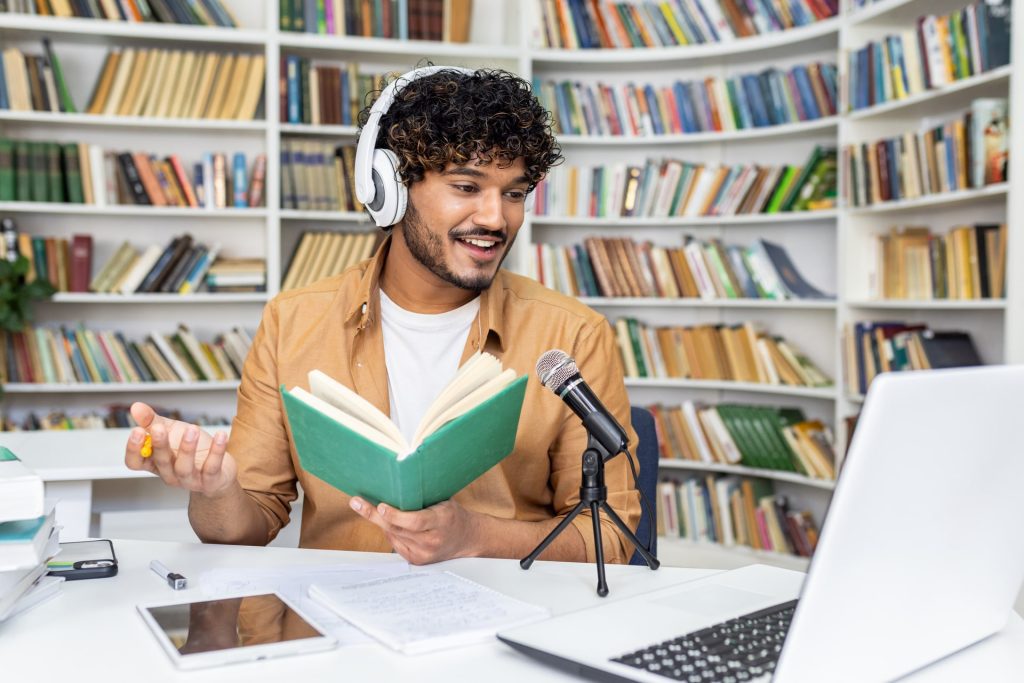 A young male podcaster records in a cozy home studio set against a bookshelf backdrop, evoking a vibe of knowledge and creativity. His engagement with audience is palpable even through visual medium.
