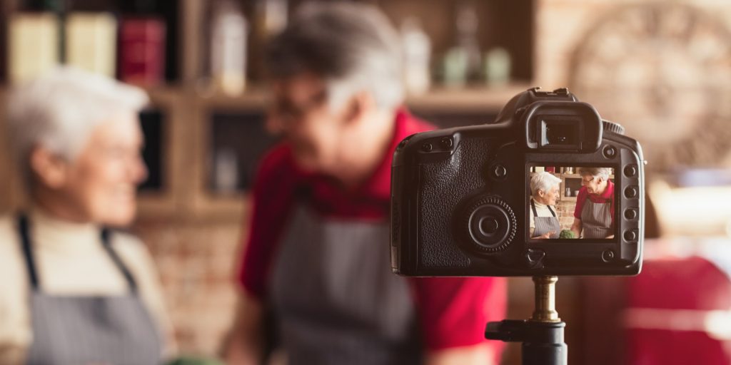 A happy senior couple, wearing aprons, stand in a home kitchen and film a cooking video. The couple smiles as they look at the camera, which is mounted on a tripod.
