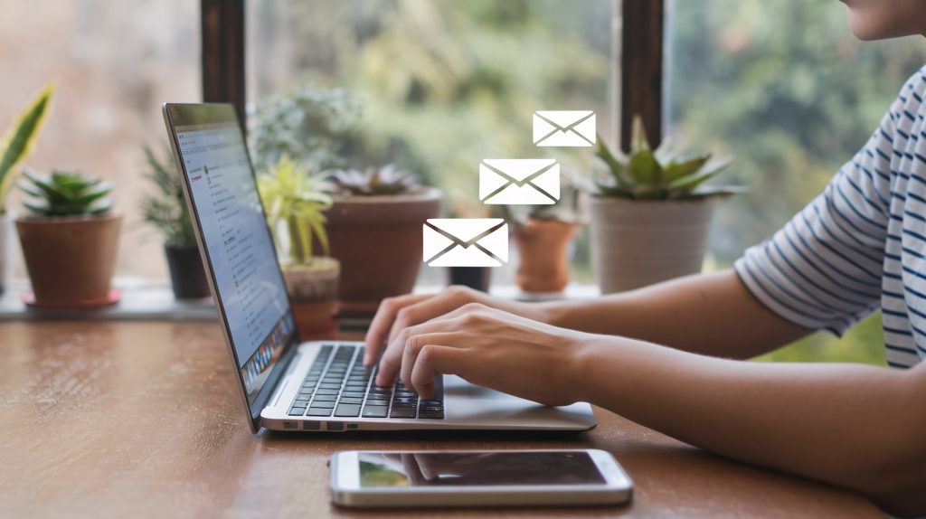 A person typing on a laptop at a wooden desk surrounded by potted plants, with email icons above.