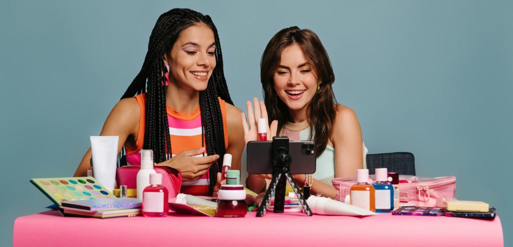 Two confident young woman recording make-up tutorial while sitting at the desk against blue background.
