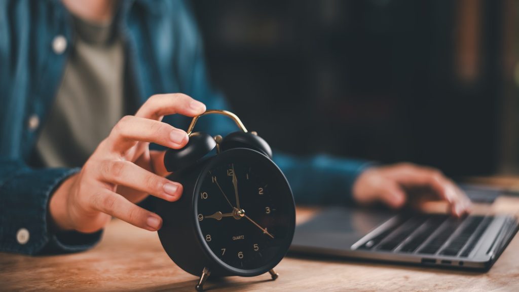 Hand holding alarm clock beside laptop showing concept of deadline, time control, efficiency, and business productivity for workplace, study, planning, and remote working lifestyle.
